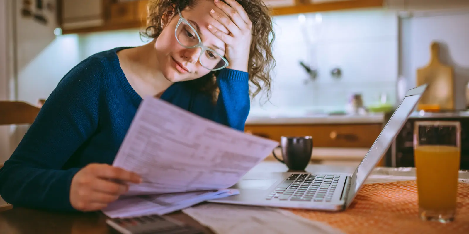 A woman in glasses looking worried as she reads a paper bill at a kitchen table with a laptop and calculator