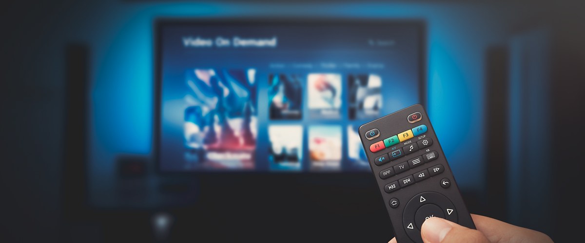 A hand holding a remote control in front of a TV showing a video-on-demand menu