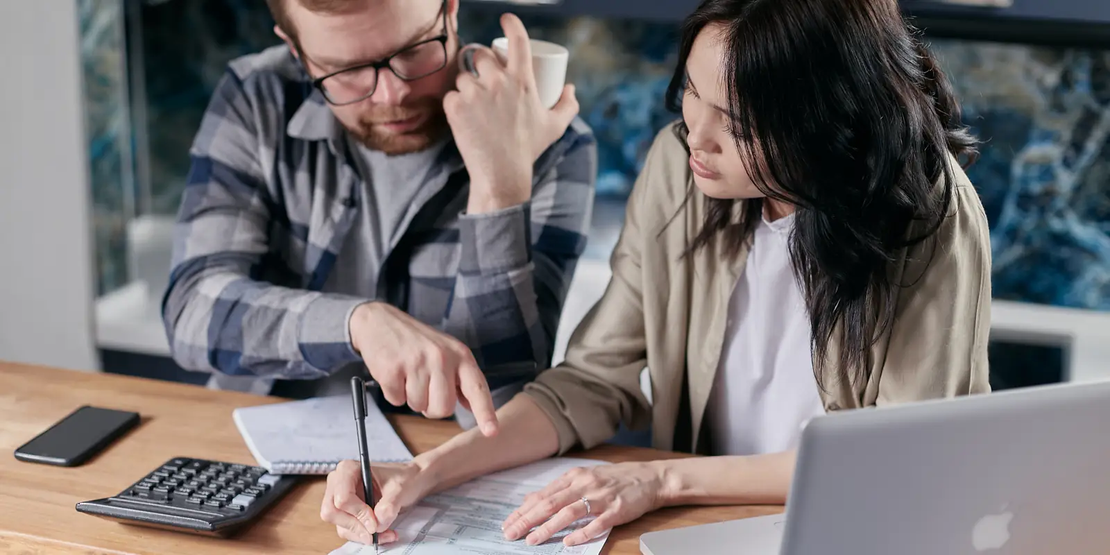 A man and a woman at a kitchen table going through a bill together with a calculator, notebook and laptop